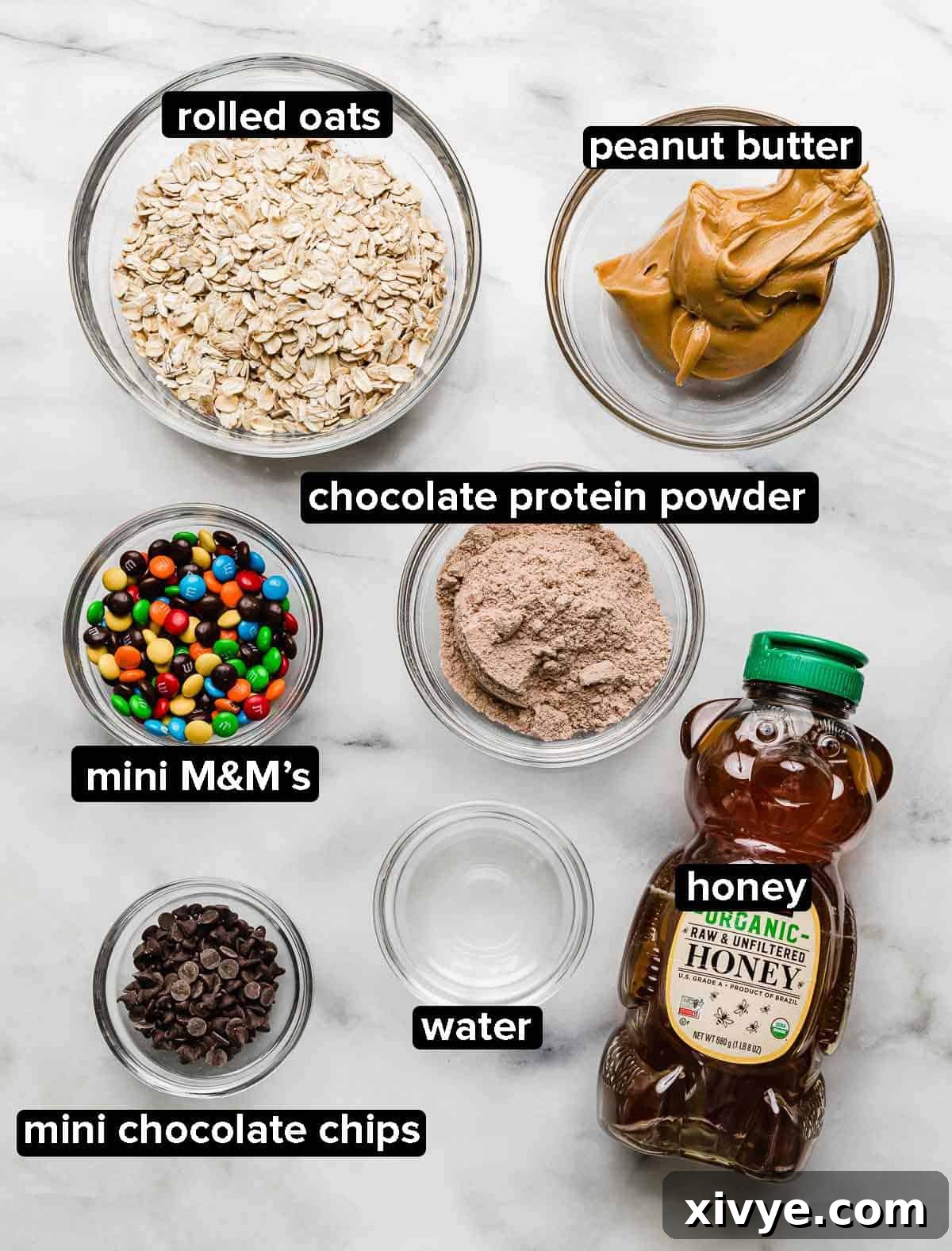 All the essential Monster Cookie Protein Balls recipe ingredients laid out neatly on a white background, including old-fashioned oats, creamy peanut butter, golden honey, chocolate protein powder, vibrant mini M&M's, clear water, and delightful mini chocolate chips, ready for assembly.