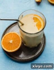 A thick Orange Julius drink recipe in a glass cup with an orange slice in the drink, on a blue background.
