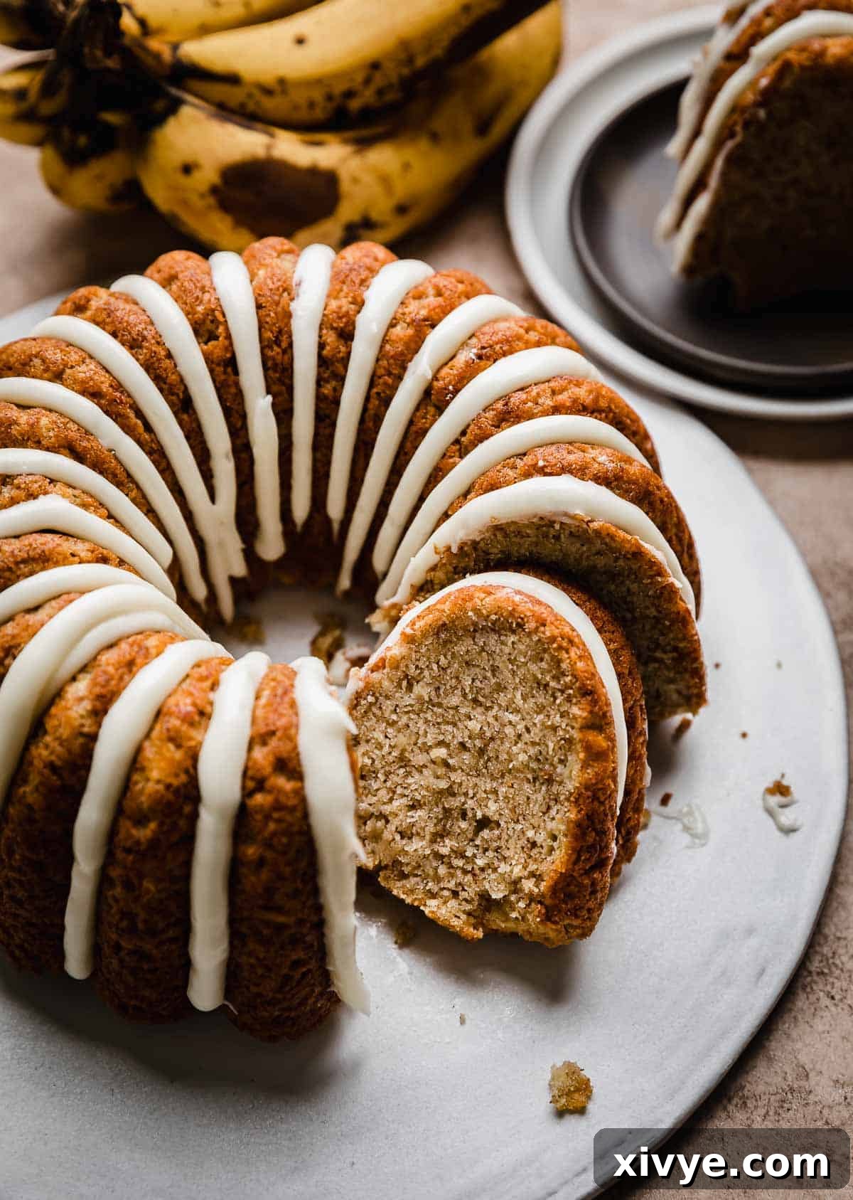 Golden Banana Bundt Delight 2 A glazed Banana Bread Bundt Cake on a white cake plate with a slice of banana bundt cake tilting against the full cake.