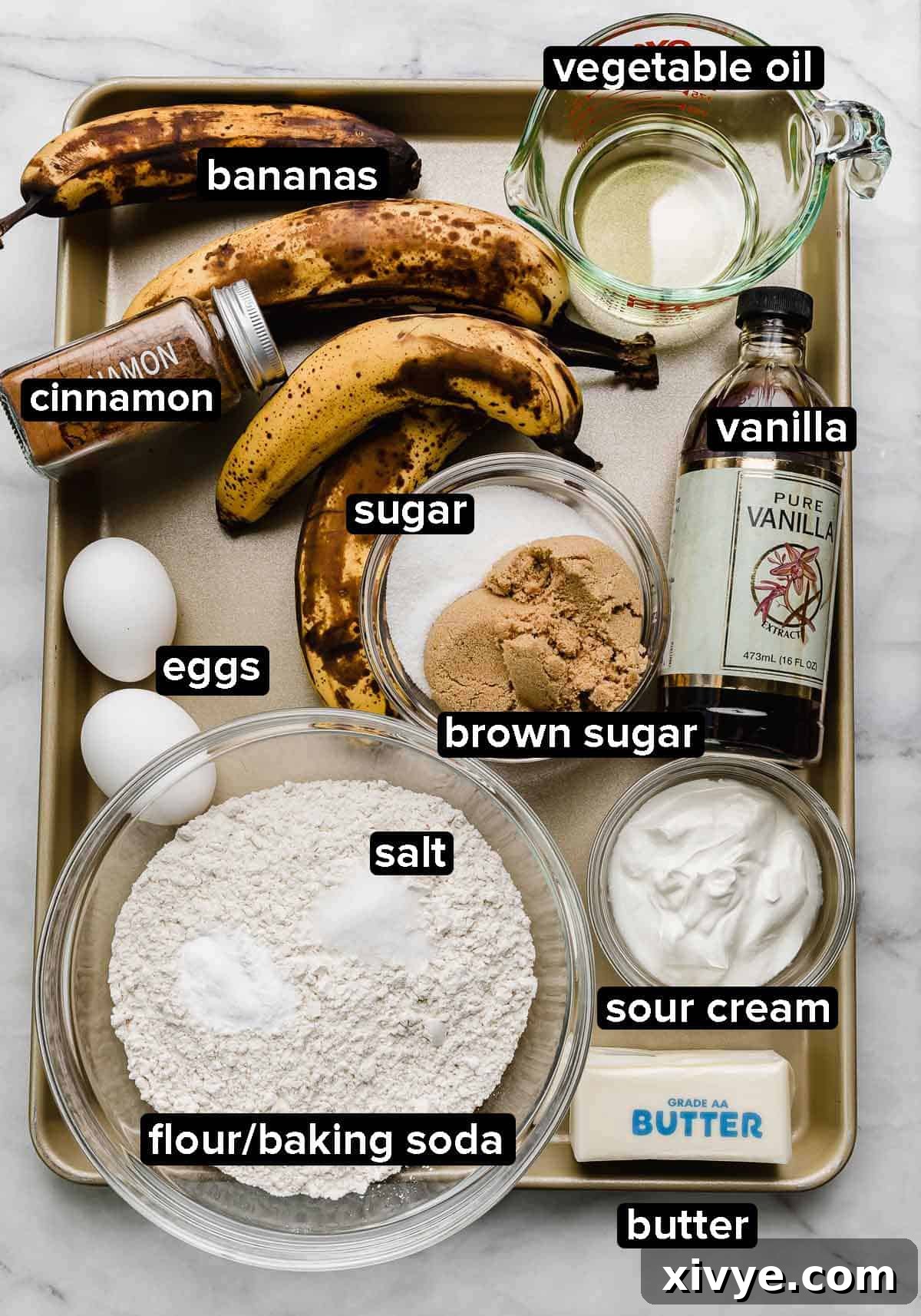 Golden Banana Bundt Delight 3 Banana Bread Bundt Cake ingredients on a gold baking sheet: overly ripe bananas, sour cream, butter, eggs, oil, vanilla, sugars, and other dry ingredients.