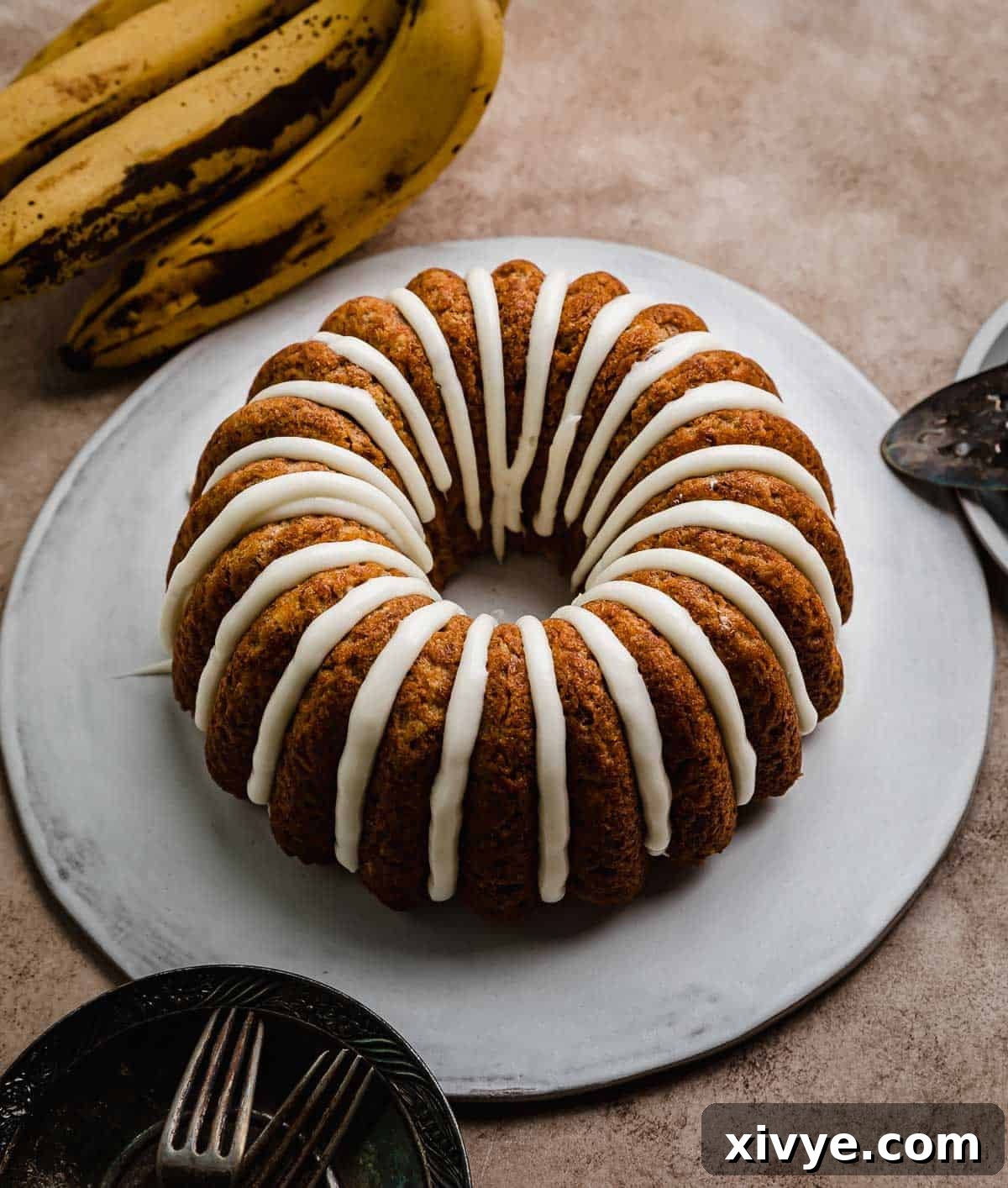 Golden Banana Bundt Delight 7 A cream cheese glazed banana bundt cake on a white cake plate that's on a brown textured background.