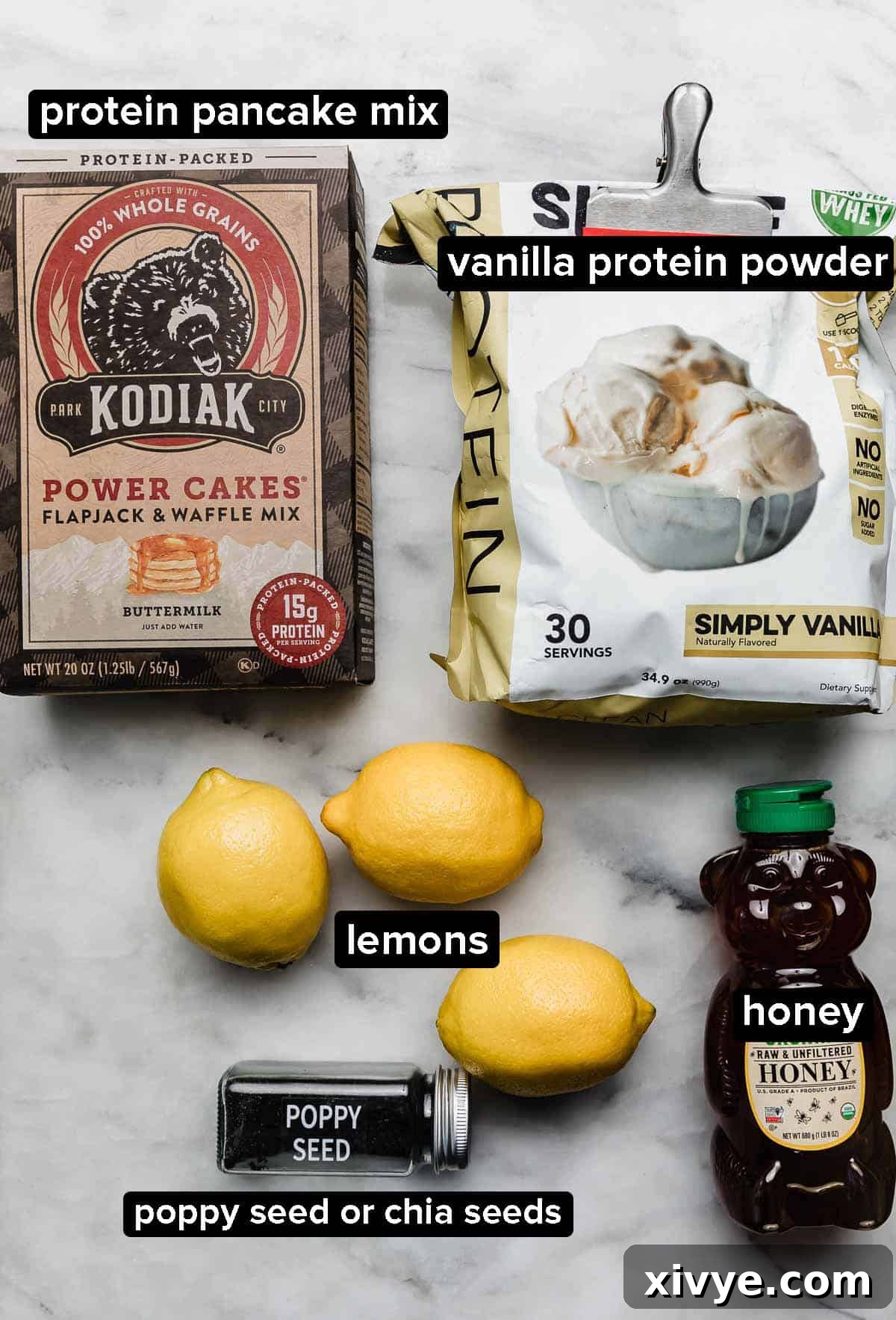 Lemon Protein Ball ingredients on a white marble surface: vanilla protein powder, lemons, honey, protein pancake mix, and poppy seeds.