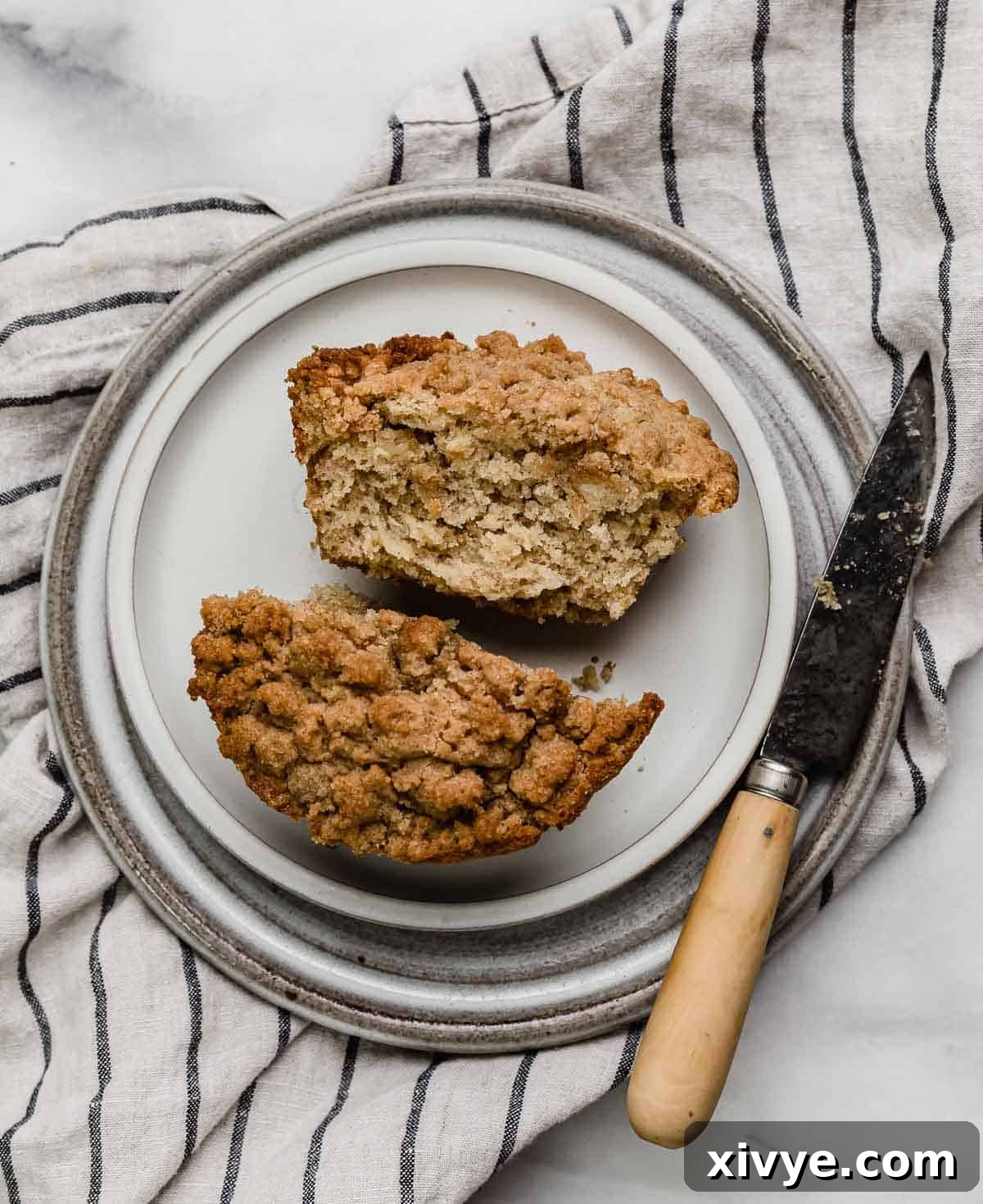 Overhead photo of a Banana Bread Crumb Muffin cut in half on a white plate with a knife to the right of the muffin.