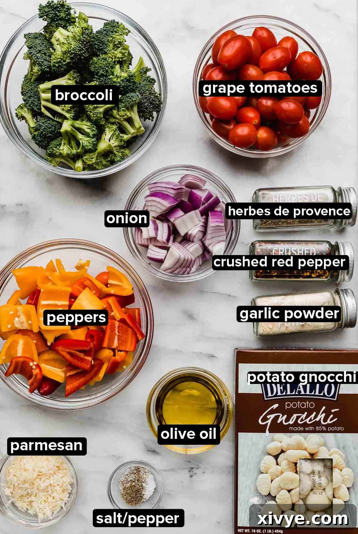 Individual ingredients for Sheet Pan Gnocchi and Vegetables displayed in glass bowls: uncooked gnocchi, vibrant broccoli, mixed baby bell peppers, sliced red onion, grape tomatoes, grated Parmesan cheese, and a bottle of olive oil, all arranged on a white marble surface.