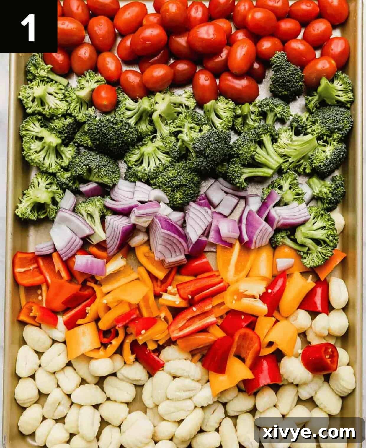 Raw gnocchi and various chopped vegetables including red onion, bell peppers, broccoli florets, and grape tomatoes, arranged in neat sections on a large baking sheet before seasoning.