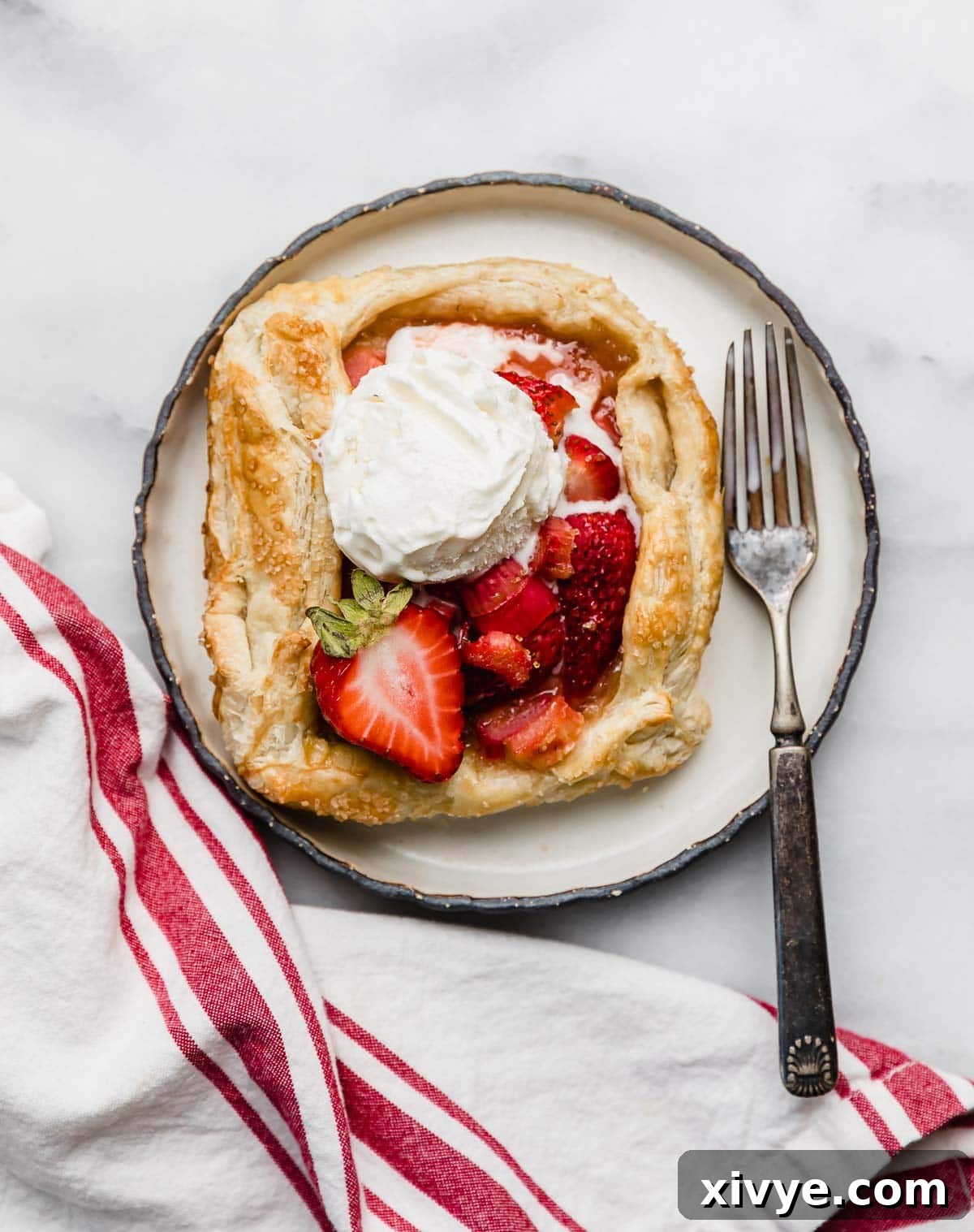 Sweet and Tart Rhubarb Strawberry Galette 7 A scoop of vanilla ice cream a top a strawberry and rhubarb galette recipe on a black lined plate on a white marble surface.