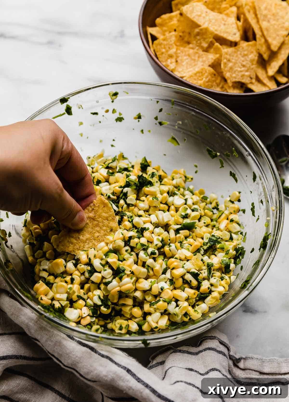 A hand holding a tortilla chip, scooping up some corn salsa with jalapeño and cilantro.