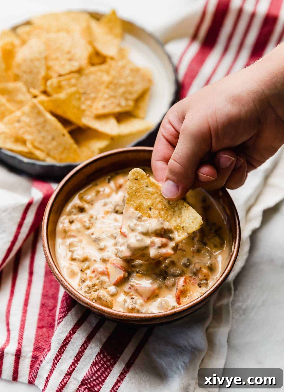 Kickin' Cream Cheese Sausage Dip 2 A brown bowl filled with Cream Cheese Sausage Rotel Dip and a hand scooping a tortilla chip into the dip.