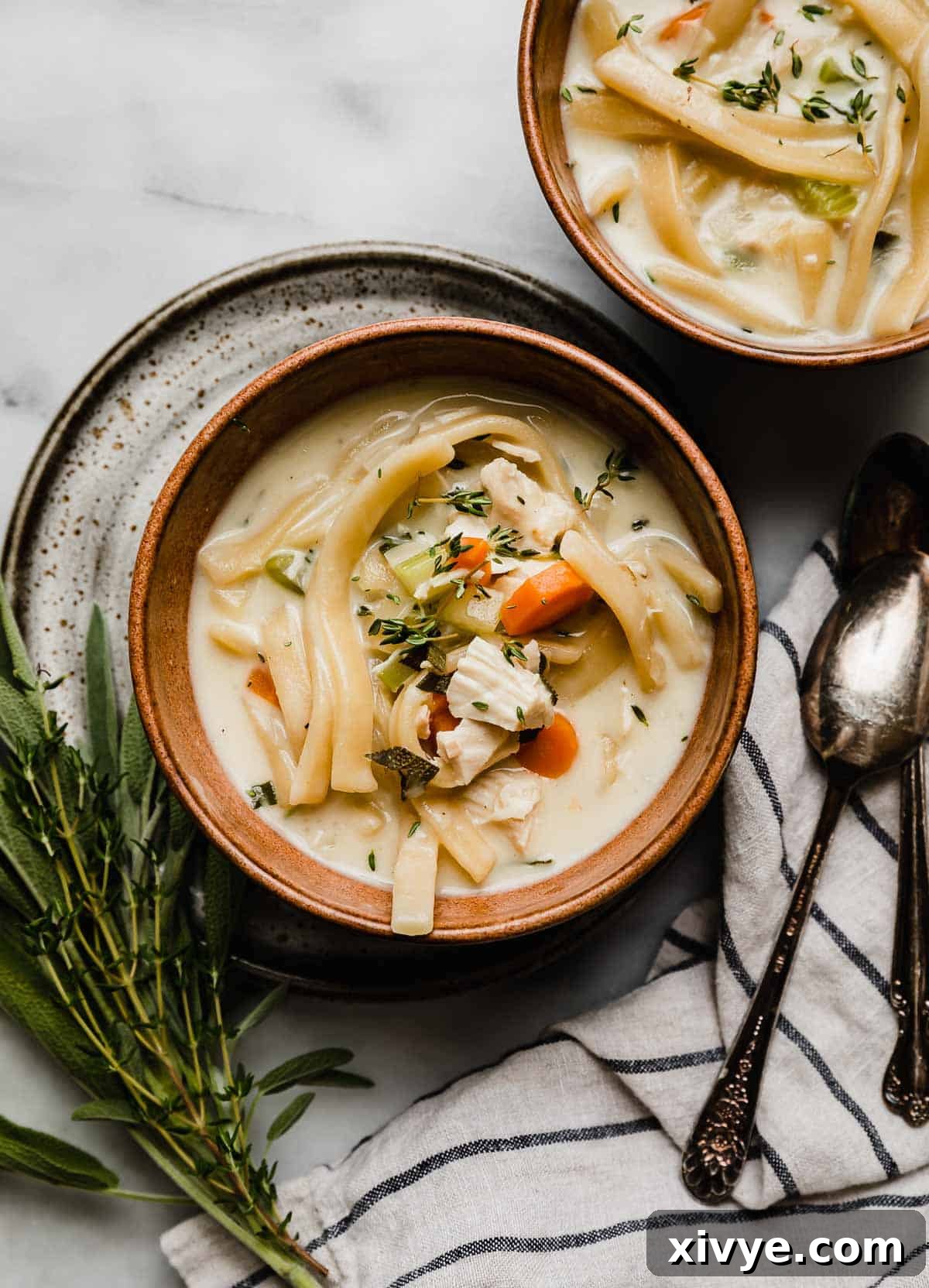 A bowl of creamy chicken noodle soup, generously garnished with fresh herbs, resting on a pristine white marble surface. The golden broth and plump noodles invite a comforting meal.