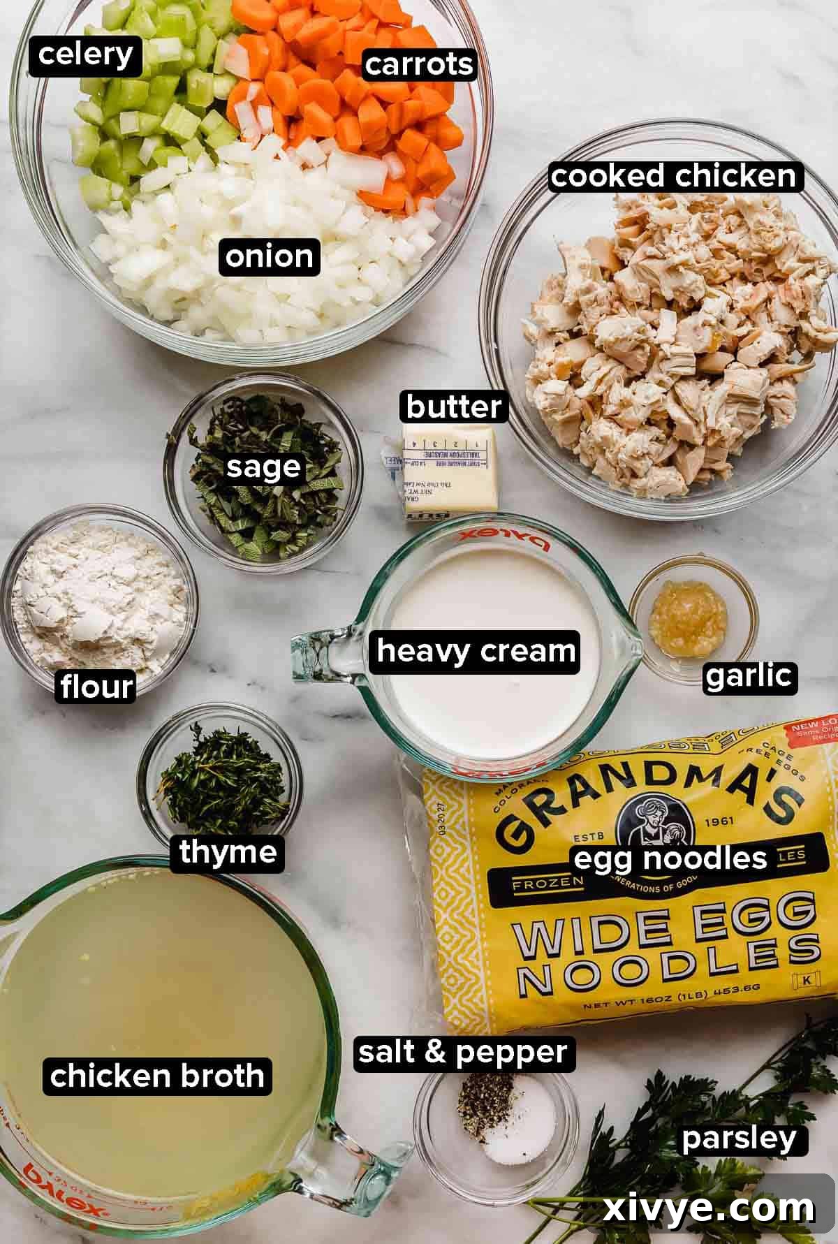 A vibrant display of fresh and prepared ingredients for creamy chicken noodle soup on a white marble surface, including chopped vegetables, cooked chicken, fresh herbs, wide egg noodles, broth, butter, flour, and heavy cream.