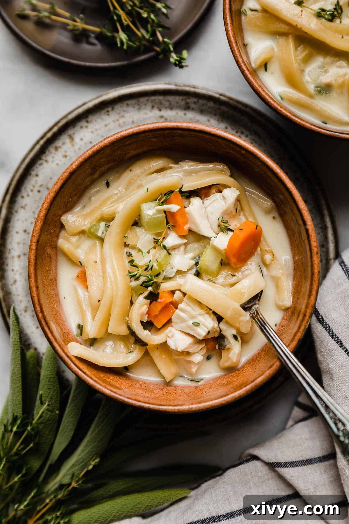 A close-up view of the rich, creamy chicken noodle soup, beautifully garnished with fresh thyme, presented in an inviting brown bowl, highlighting its comforting textures and colors.