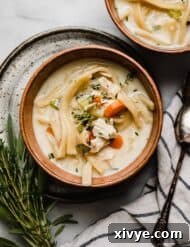 Creamy Chicken Noodle Soup in a brown colored bowl on a white marble surface with fresh herbs to the left of the bowl.