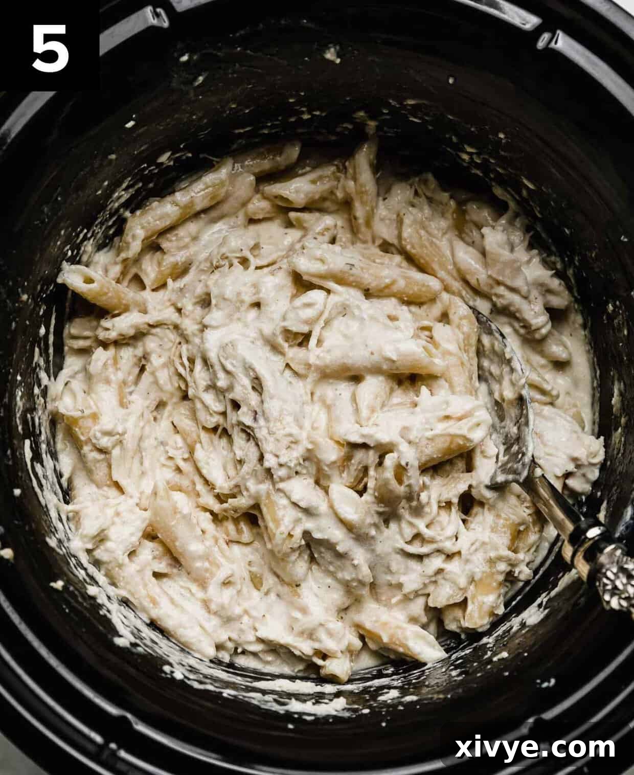 A chicken Alfredo recipe made in a black crock pot with a spoon scooping into the creamy shredded chicken Alfredo mixture.