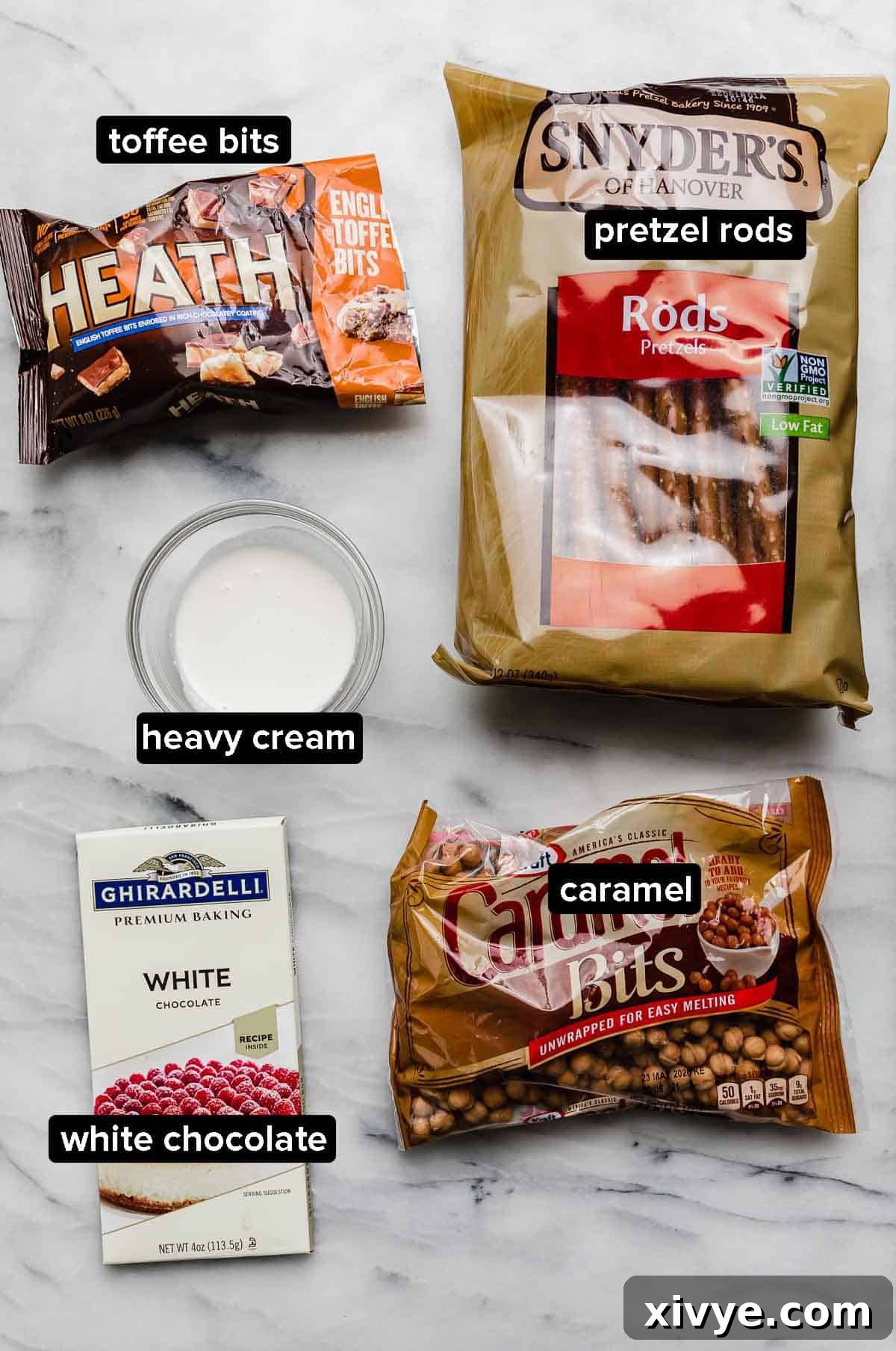 Toffee Caramel dipped pretzels ingredients on a white marble surface: pretzel rods, caramel, white chocolate, heavy cream, and heath toffee bits. 