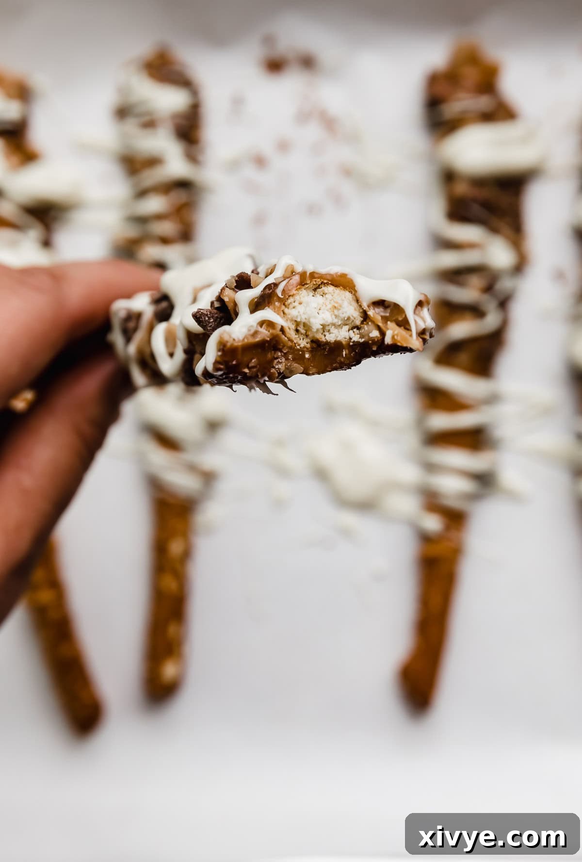 A hand holding a caramel dipped pretzel rod rolled in toffee bits and white chocolate, with a bite taken out of the fancy pretzel.