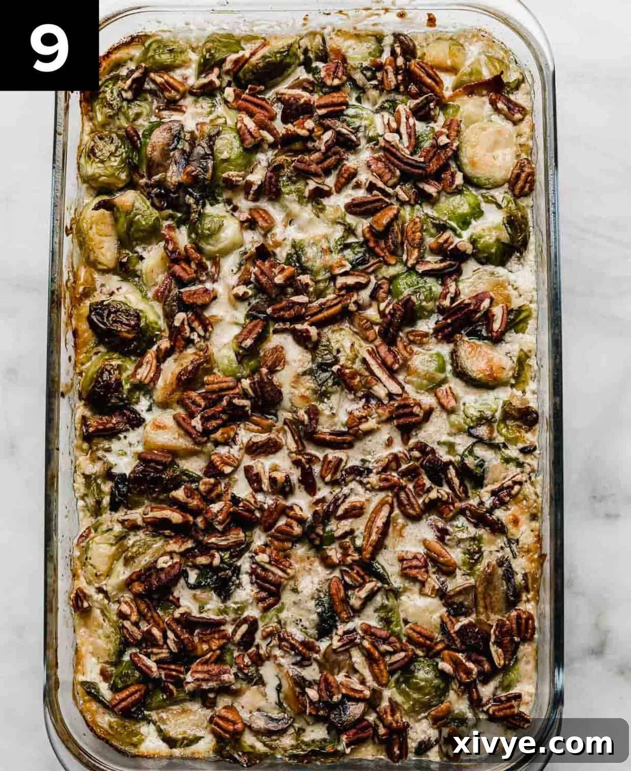 Baked Brussels Sprouts Gratin with Bacon and gruyere and heavy cream mixture, topped with chopped pecans in a glass rectangle pan on a white marble surface.