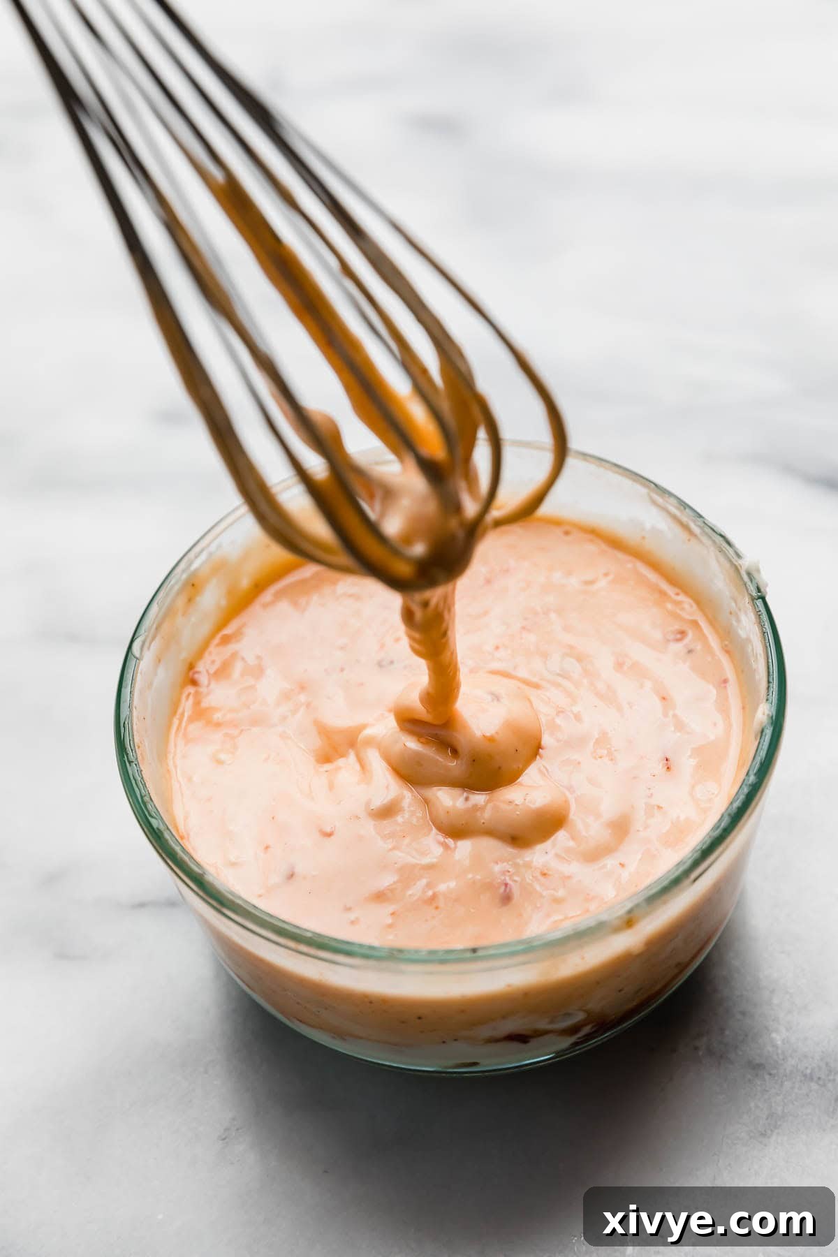A whisk drizzling a creamy bang bang sauce into a glass bowl.