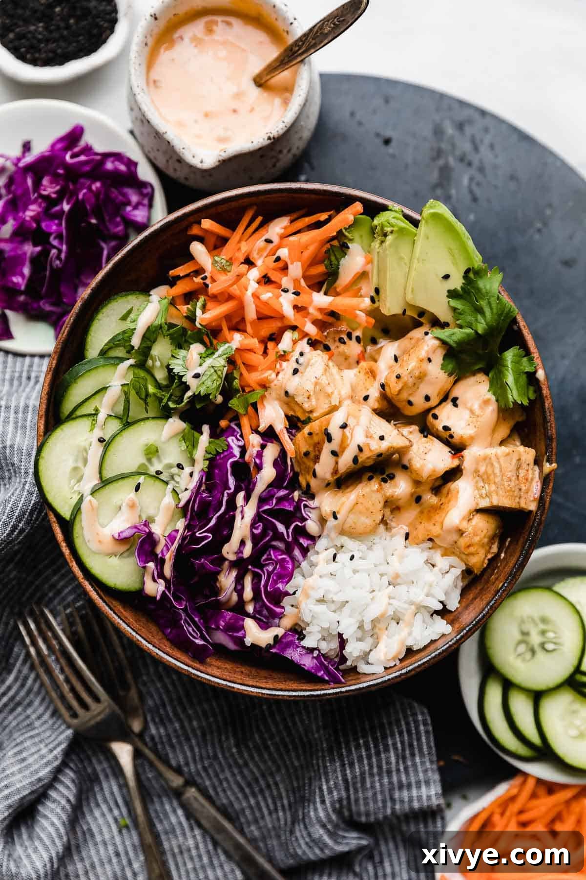 Effortless Bang Bang Chicken Bowls 2 A brown bowl filled with the best bang bang chicken bowl recipe with cooked rice, red cabbage, sliced cucumbers, shredded carrots, avocado, and bang bang sauce drizzled overtop.