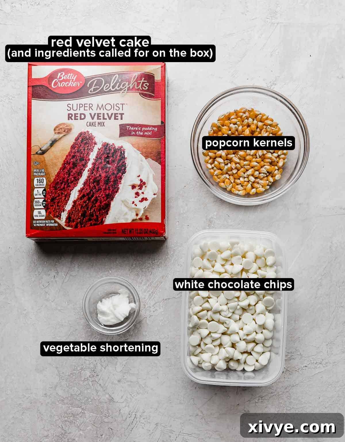 A flat lay showing Red Velvet Popcorn ingredients on a light gray background: red velvet cake mix box, popcorn kernels, white chocolate chips, and a small dish of vegetable shortening.
