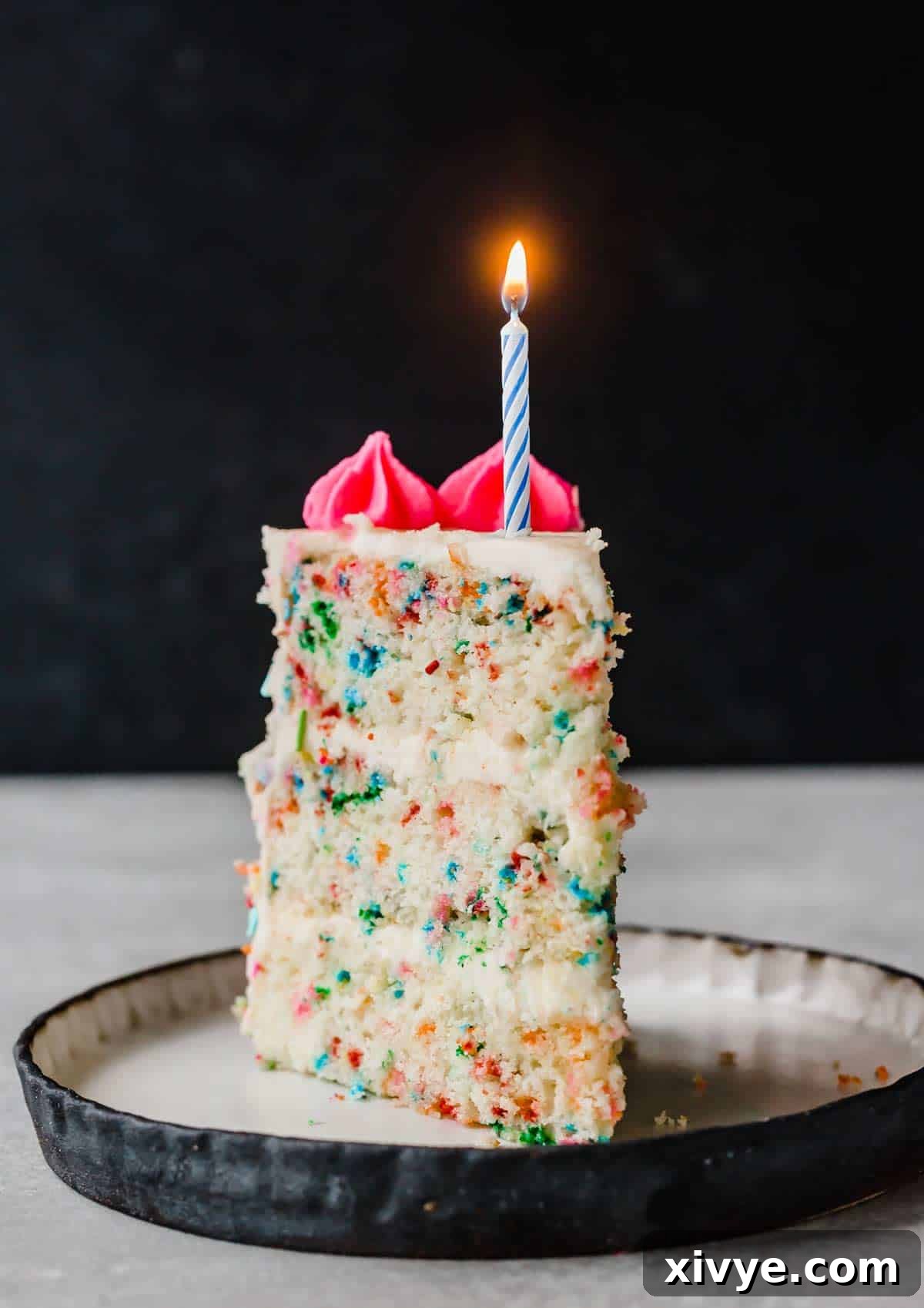 A beautifully decorated slice of Funfetti Cake on a white plate, topped with a lit blue birthday candle, showcasing the colorful sprinkles within the cake layers.