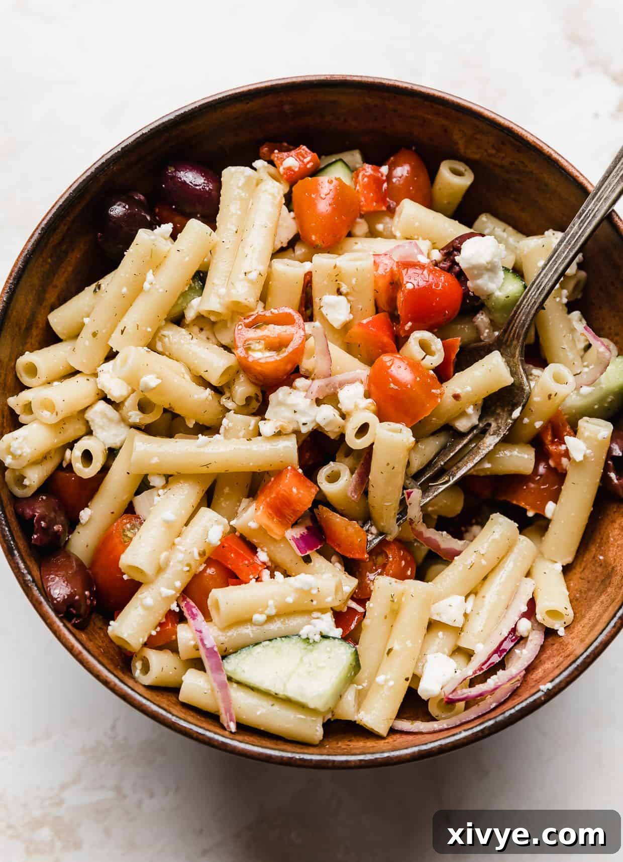 Zesty Mediterranean Pasta Salad 2 A brown bowl filled with vibrant Fresh Greek Pasta Salad, showcasing penne pasta, various chopped vegetables, and crumbled feta cheese.