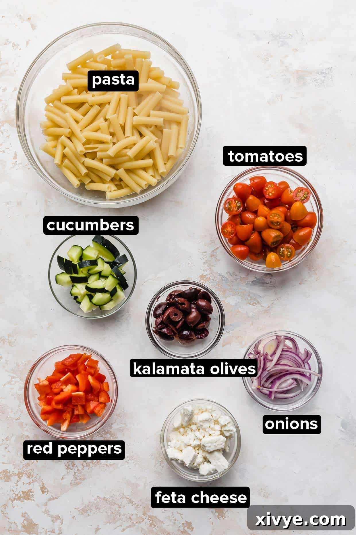Zesty Mediterranean Pasta Salad 3 A beautiful arrangement of fresh ingredients including ripe tomatoes, cucumbers, red bell pepper, red onion, Kalamata olives, feta cheese, and dry pasta, laid out on a white surface.