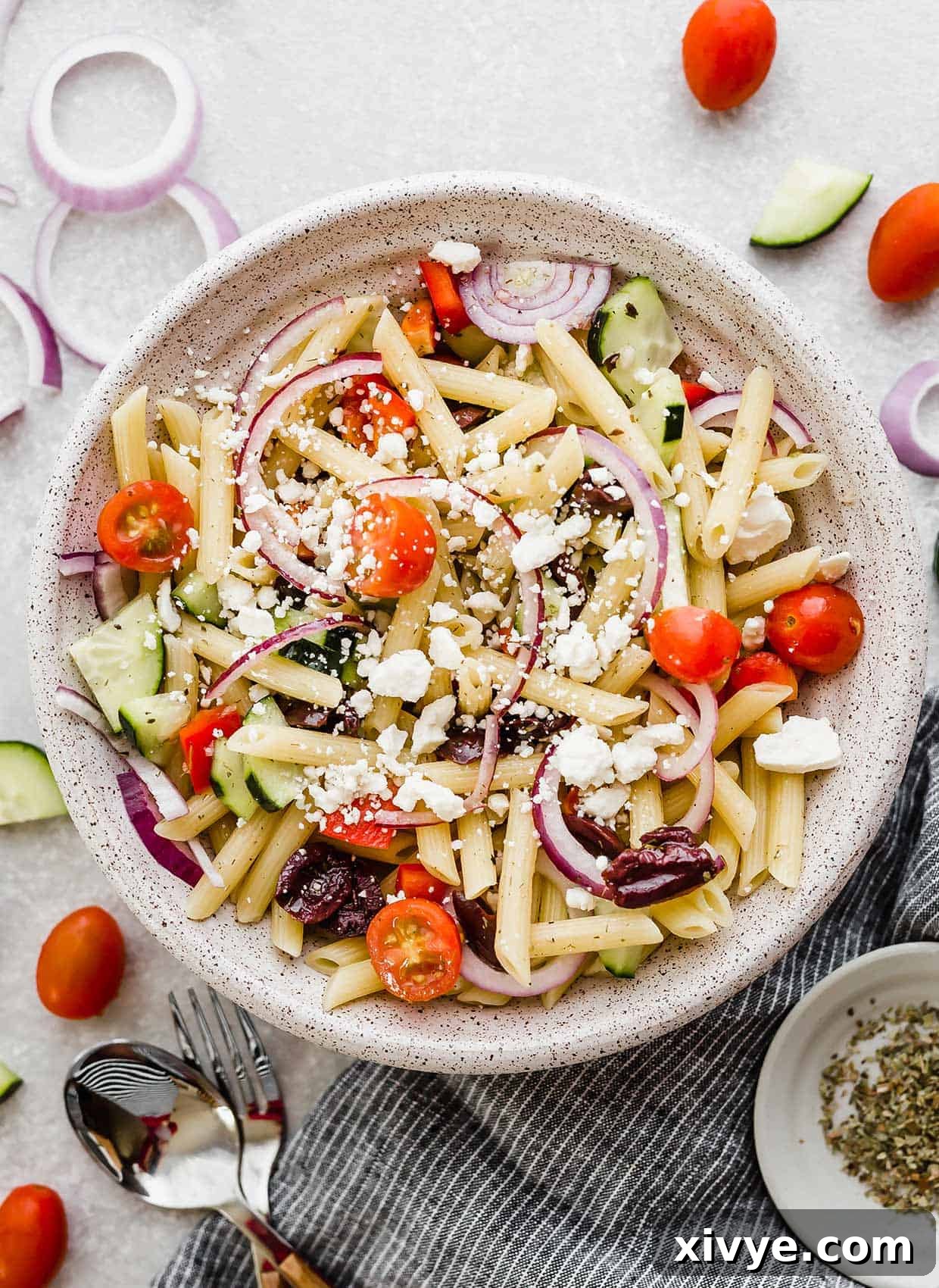 Zesty Mediterranean Pasta Salad 7 A white bowl featuring Greek pasta salad with penne pasta, surrounded by scattered cherry tomatoes and slices of red onion for garnish.