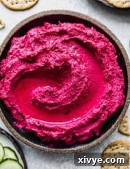Beet Hummus on a round black plate surrounded by sliced cucumbers and round whole grain crackers.