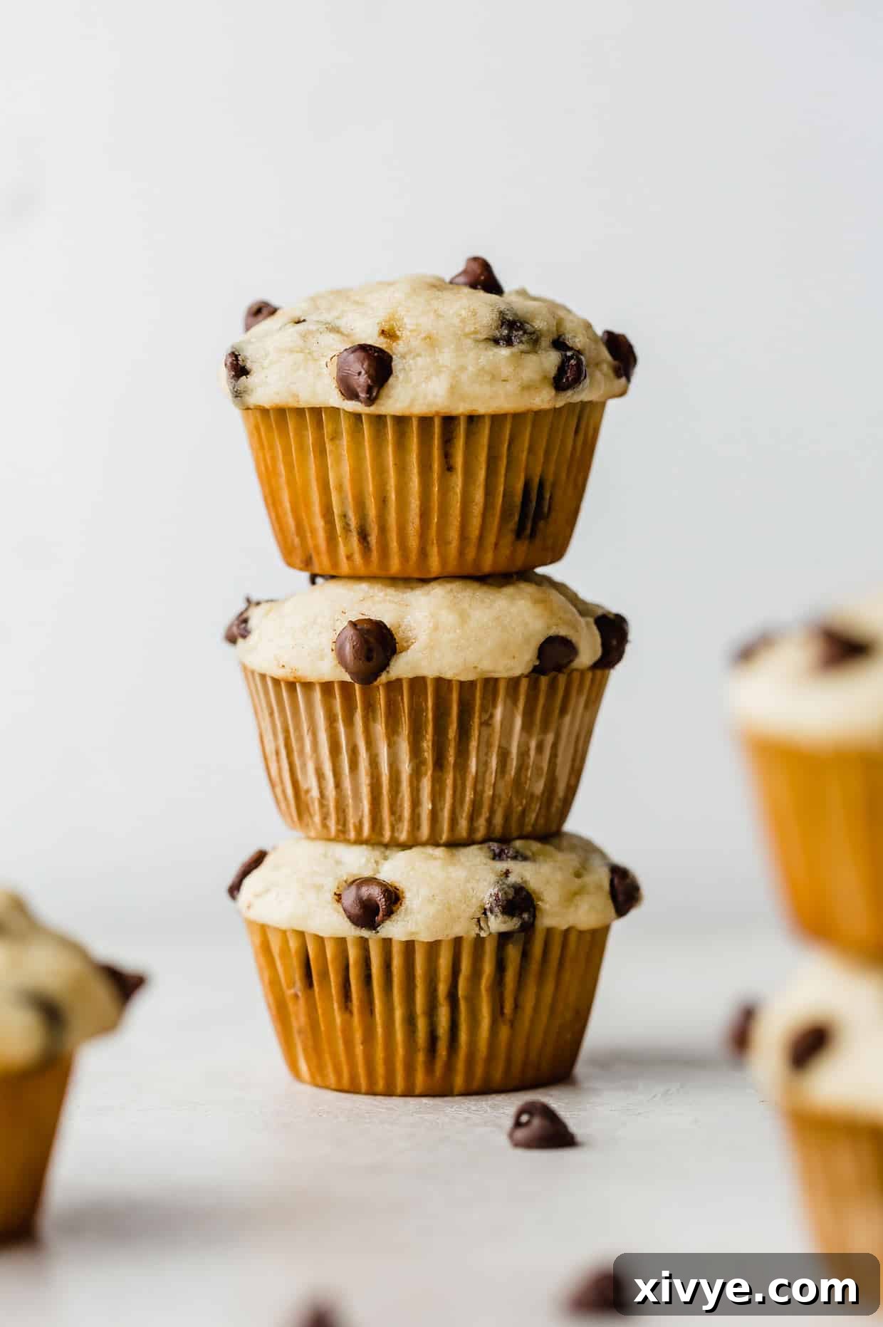 Three golden-brown buttermilk banana chocolate chip muffins, perfectly stacked on a rustic white background, showcasing their fluffy tops and visible melty chocolate chips, ready to be enjoyed.