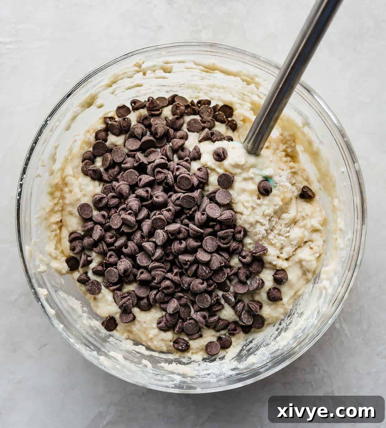 Thick, golden-brown buttermilk banana chocolate chip muffin batter, rich with banana chunks and visible chocolate chips, in a clear glass mixing bowl, ready for portioning.