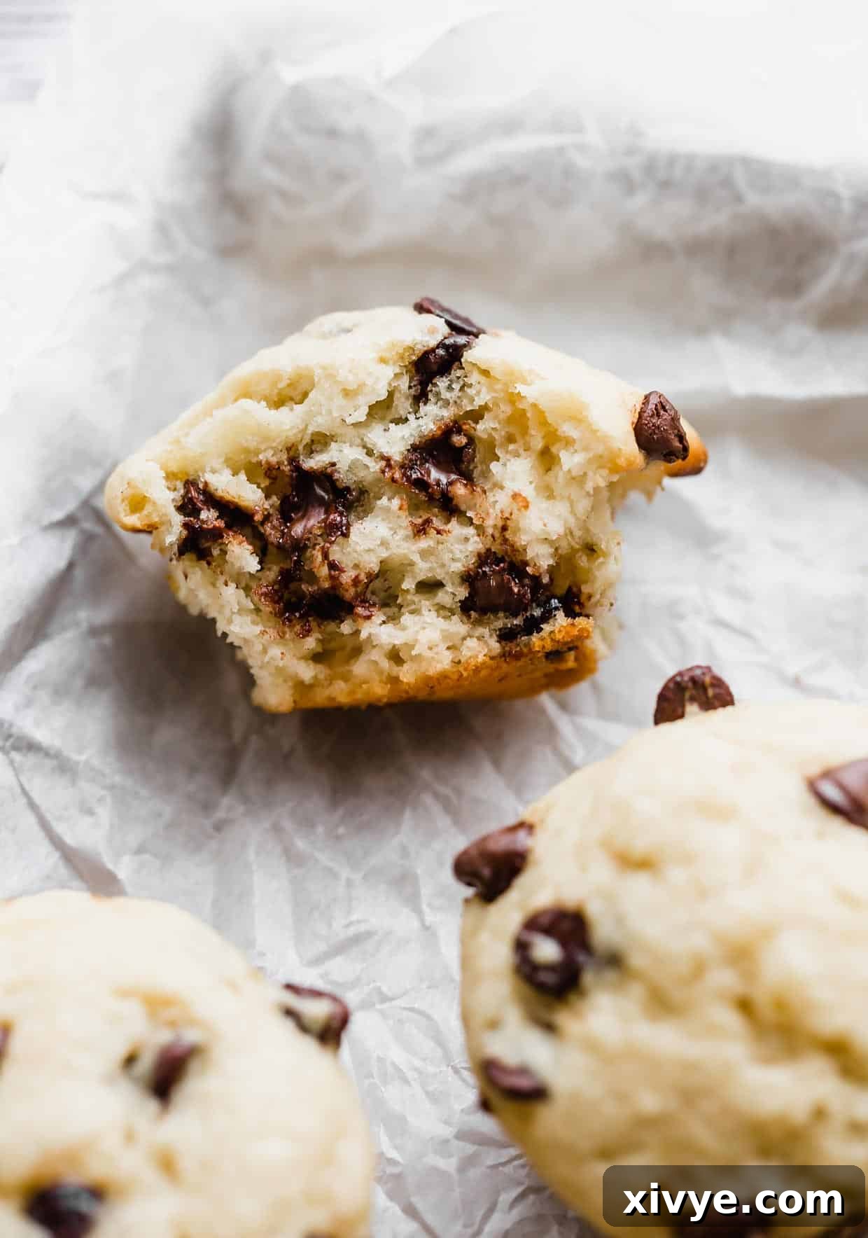 A perfectly baked buttermilk banana chocolate chip muffin, sliced in half to reveal its fluffy, moist interior and generous distribution of melted chocolate chips, resting on white parchment paper.