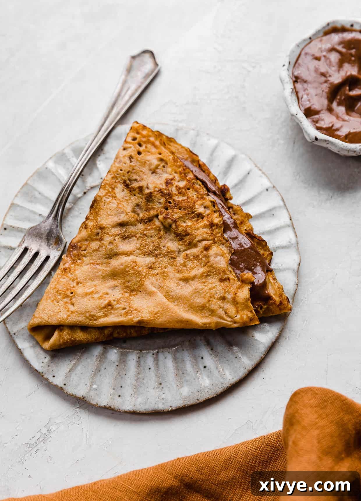 Golden Pumpkin Crepes 2 A Pumpkin Crepe filled with whipped nutella folded into a triangle, on a white plate.