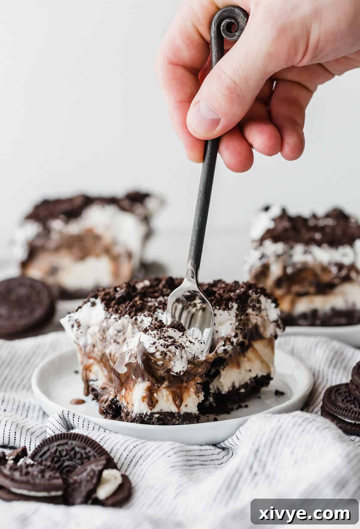Dreamy Oreo Ice Cream Cake 8 A hand stabbing a fork into the front of an Oreo Ice Cream Cake slice, on a white plate, inviting a taste.