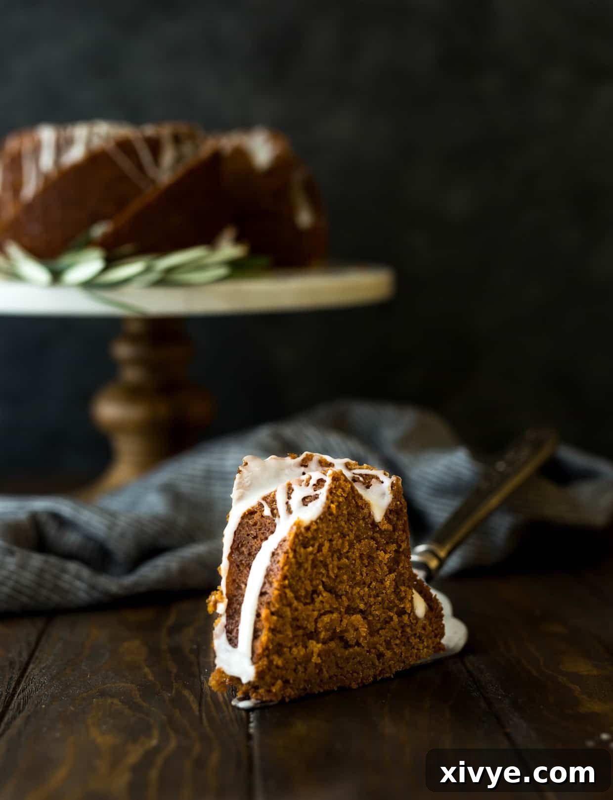 A slice of gingerbread bundt cake in the foreground and the remainder of the cake on a cake stand in the background, showcasing its delicious texture.