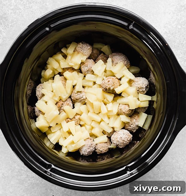 Delectable Sweet and Sour Slow Cooker Meatballs 5 A black slow cooker containing a generous portion of frozen meatballs and bright pineapple chunks, forming the base of the dish.