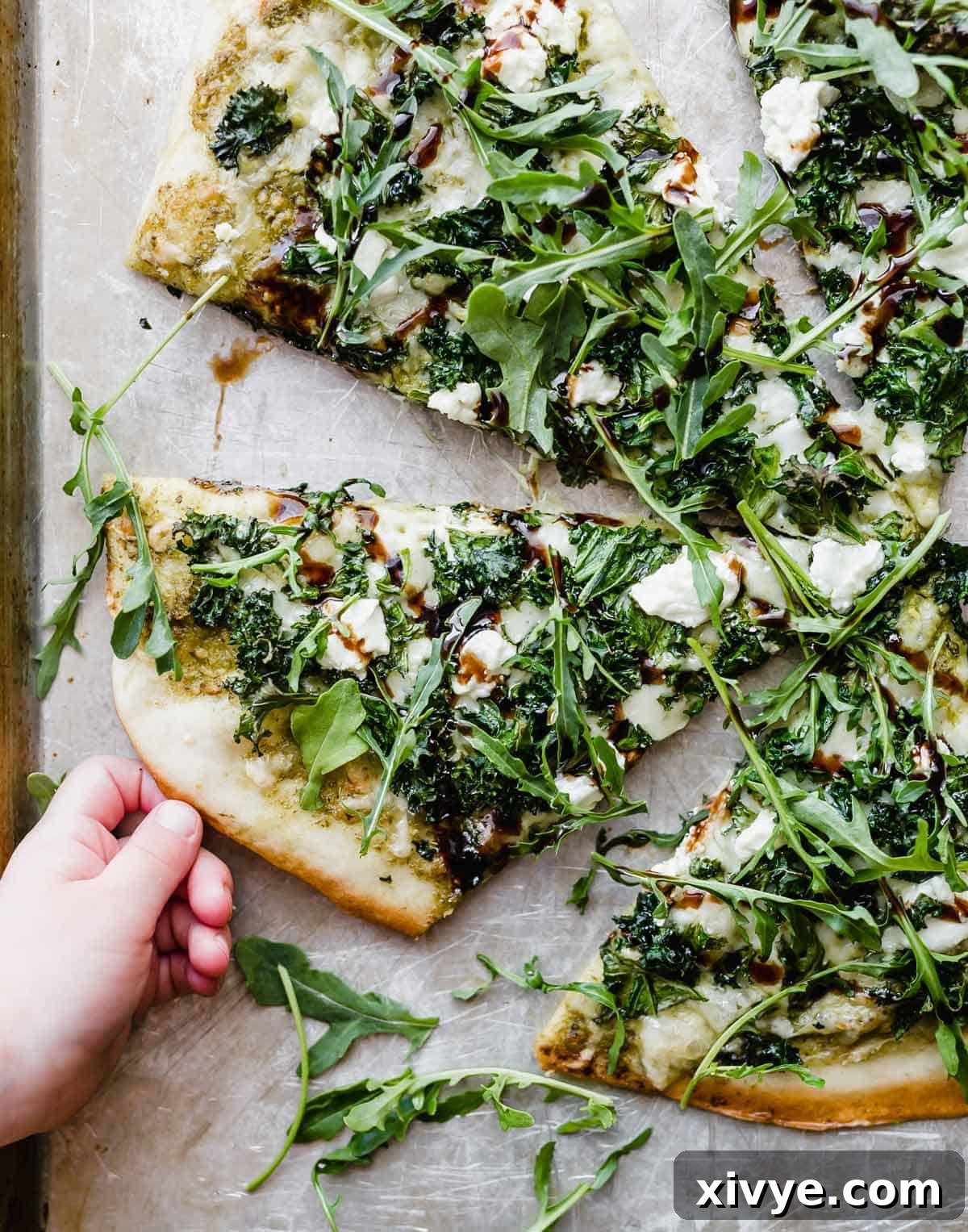 A small child's hand reaching for a slice of freshly baked homemade kale pizza, beautifully topped with fresh arugula and a drizzle of balsamic glaze.