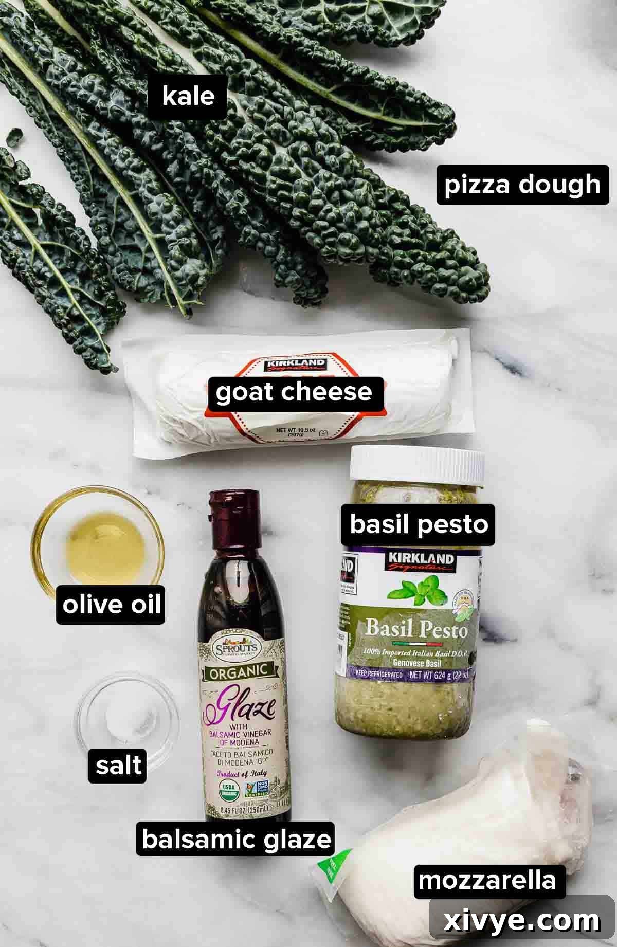 Kale Pizza ingredients displayed on a white marble surface including fresh kale, olive oil, basil pesto, crumbled goat cheese, and shredded mozzarella.