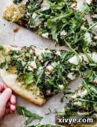 A hand grabbing a slice of kale pizza with arugula and goat cheese.