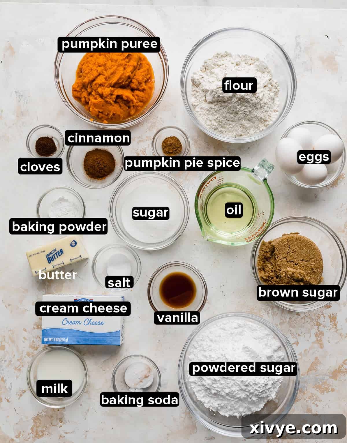 Various key ingredients for Pumpkin Layer Cake, including flour, sugars, spices, and pumpkin puree, elegantly displayed in small glass bowls on a white textured surface.