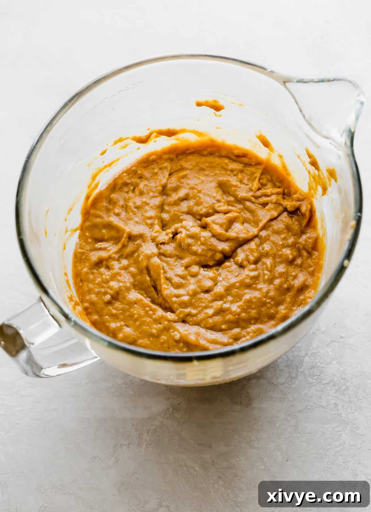 Smooth, evenly mixed pumpkin cake batter in a large glass mixing bowl, resting on a white surface, prepared for pouring into cake pans.