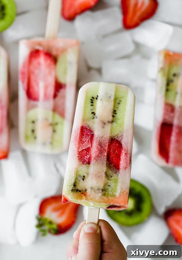 A hand holds a single kiwi strawberry popsicle, showing the vibrant fruit slices embedded within the frozen treat against a softly blurred background.