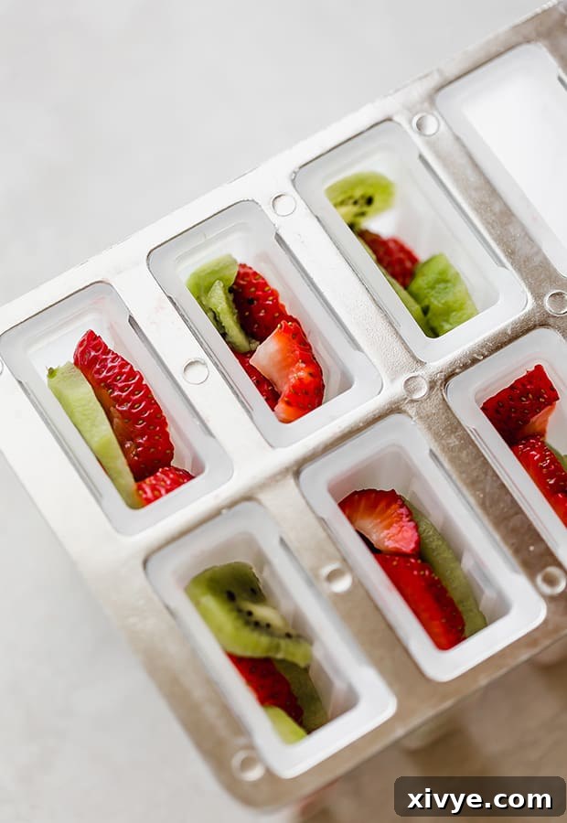 Close-up of popsicle molds neatly filled with alternating layers of thinly sliced kiwis and strawberries, ready for liquid.