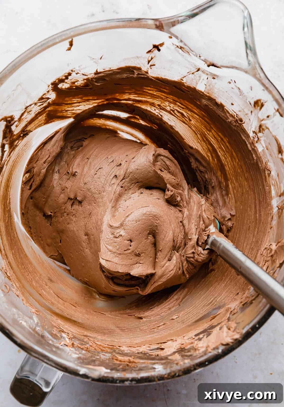 Decadent Chocolate Buttercream 13 A spatula stirring a thick, creamy, and decadent chocolate buttercream frosting made with melted chocolate, in a clear glass bowl, showcasing its rich texture.
