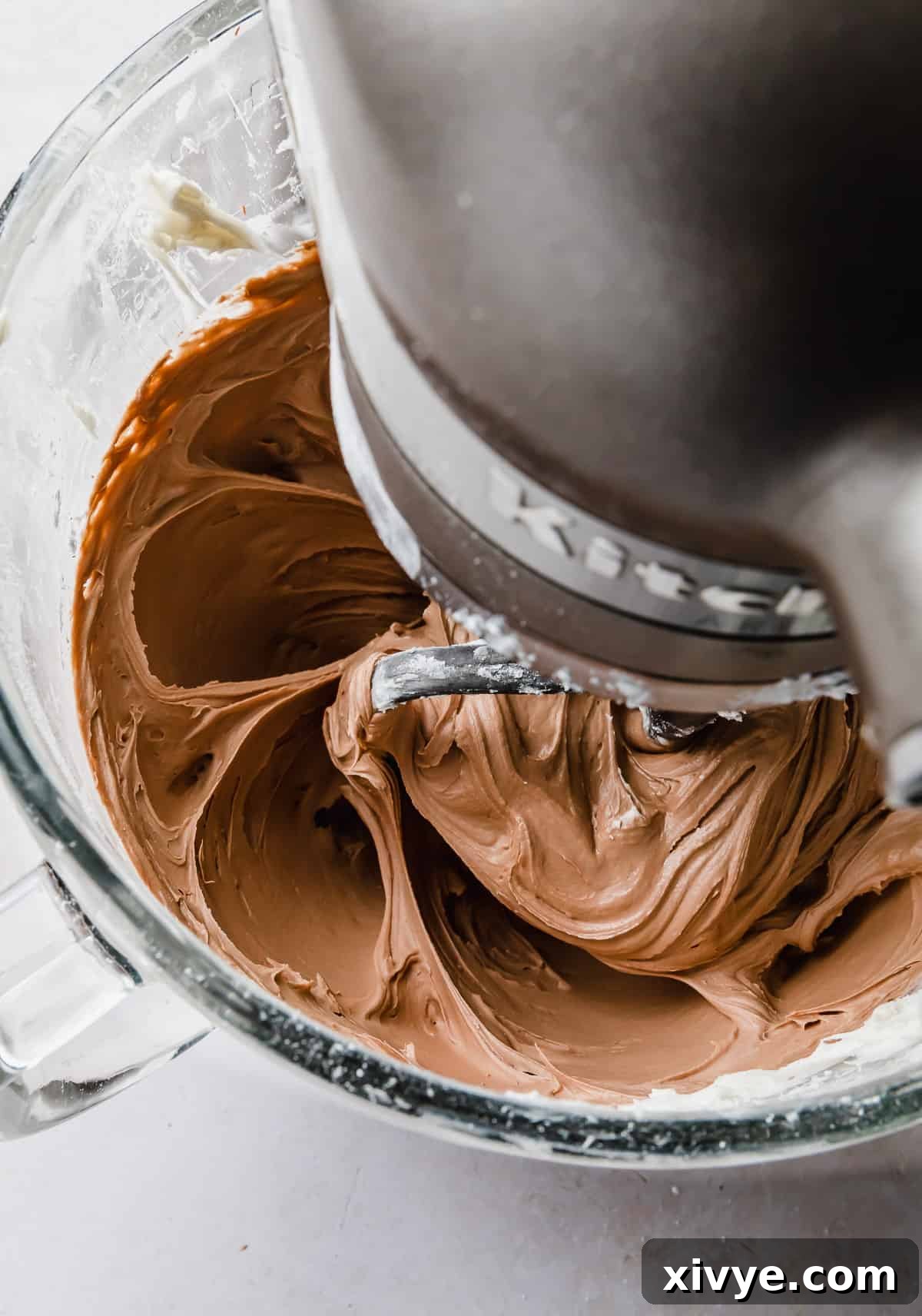 Decadent Chocolate Buttercream 10 Silky smooth Chocolate Buttercream Frosting with Melted Chocolate being vigorously stirred in a glass KitchenAid bowl, showcasing its light and fluffy consistency.