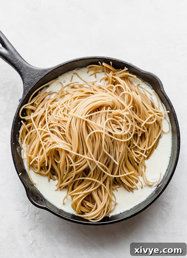 A skillet with a lemon cream sauce and freshly boiled whole wheat spaghetti noodles over the sauce.