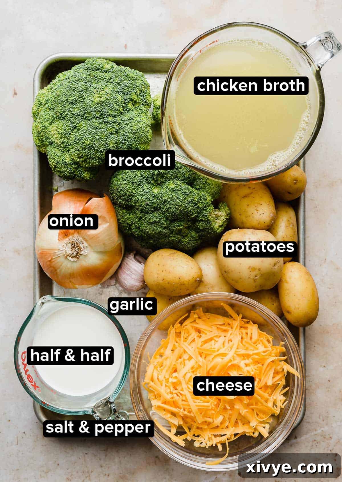 Assortment of fresh ingredients for Broccoli Potato Soup laid out on a metal baking sheet, including bright green broccoli, diced yellow onion, cubed golden potatoes, a block of cheddar cheese, a carton of half and half, chicken broth, and garlic cloves.