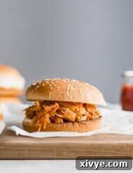 A BBQ chicken sandwich on a wooden board, with hamburger buns stacked in the background.