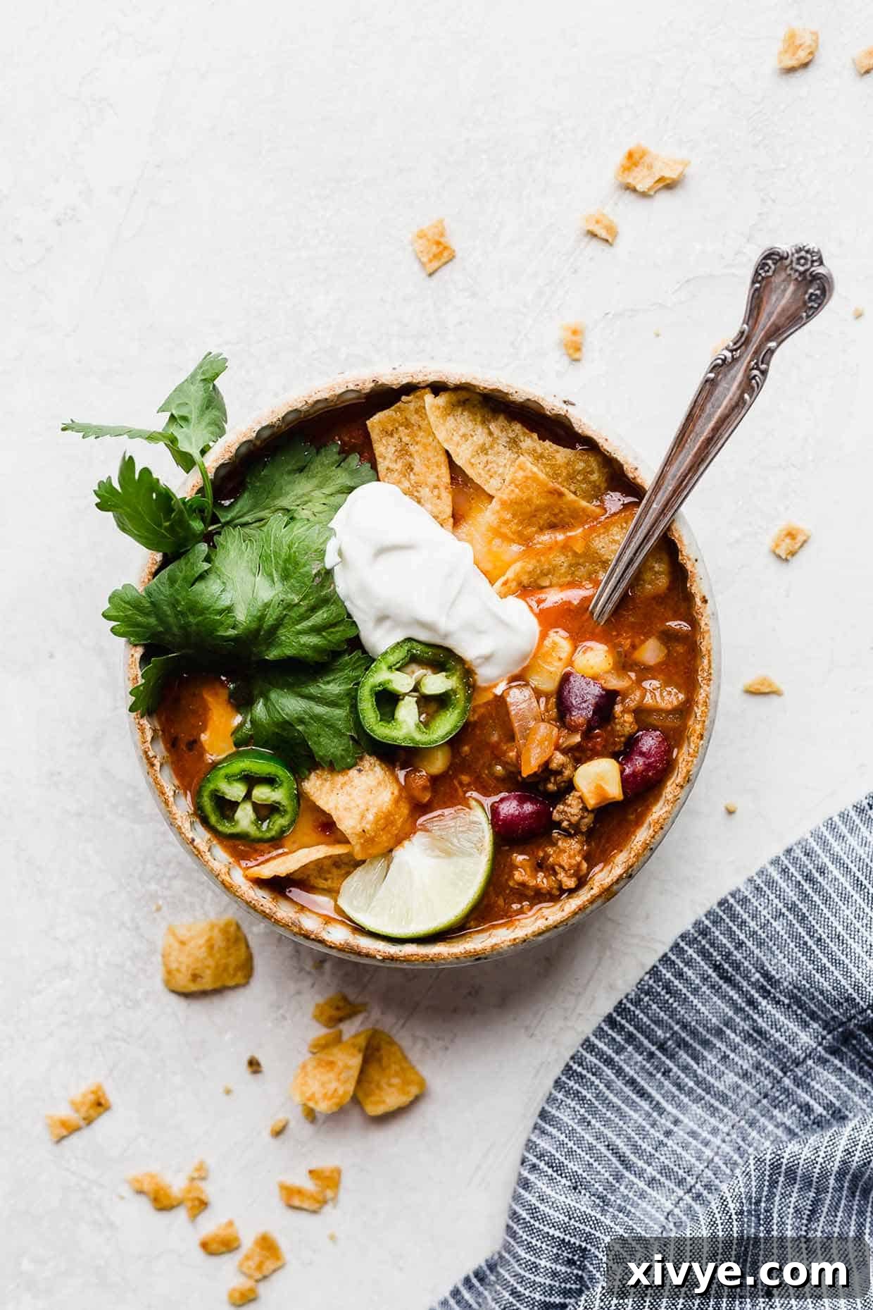 A bowl on a white background, filled with Taco Soup with Fritos, sour cream, cilantro, and sliced jalapeños.