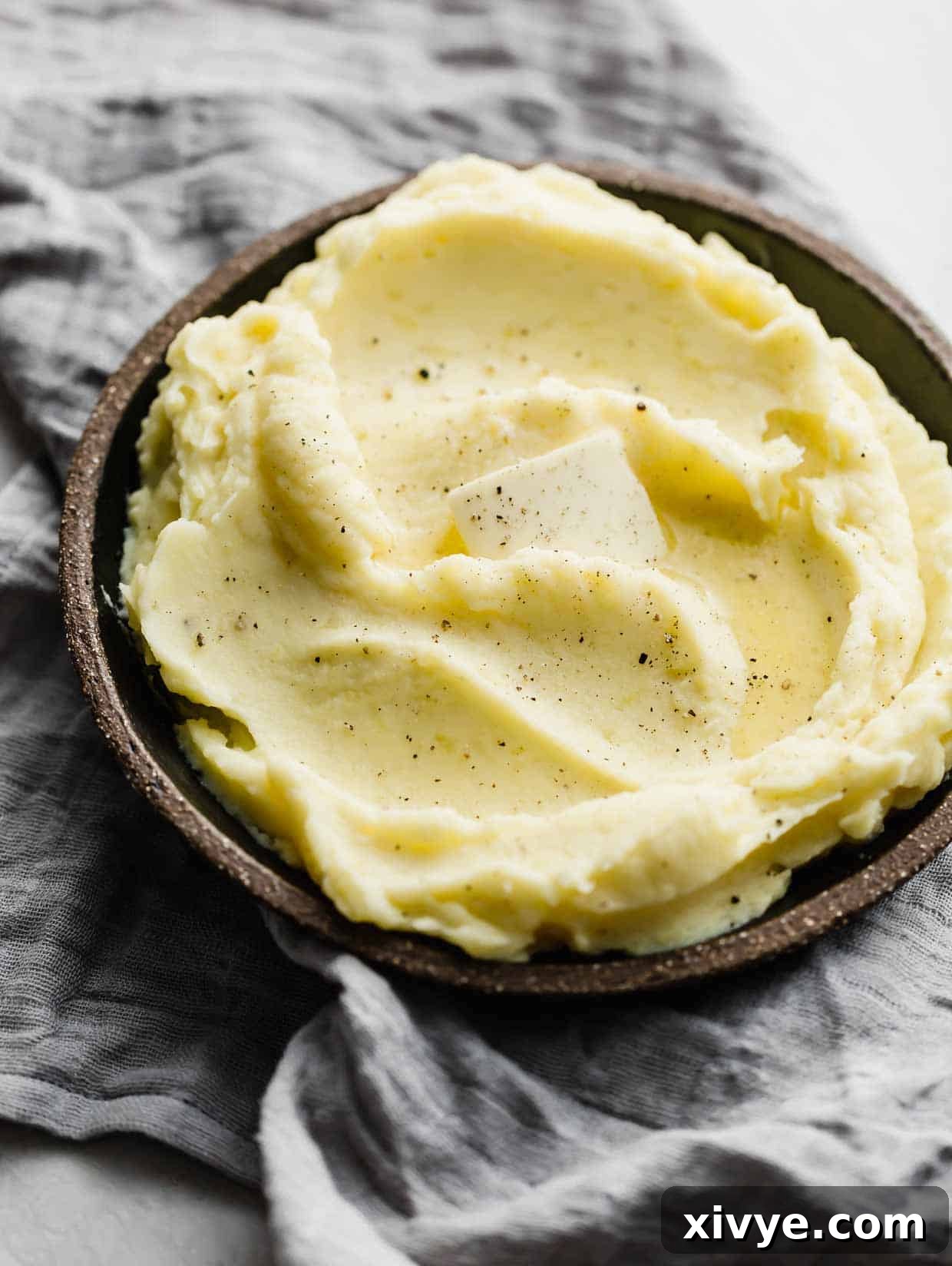 A slice of butter melting into the top of homemade mashed potatoes on a black plate, showcasing their richness.