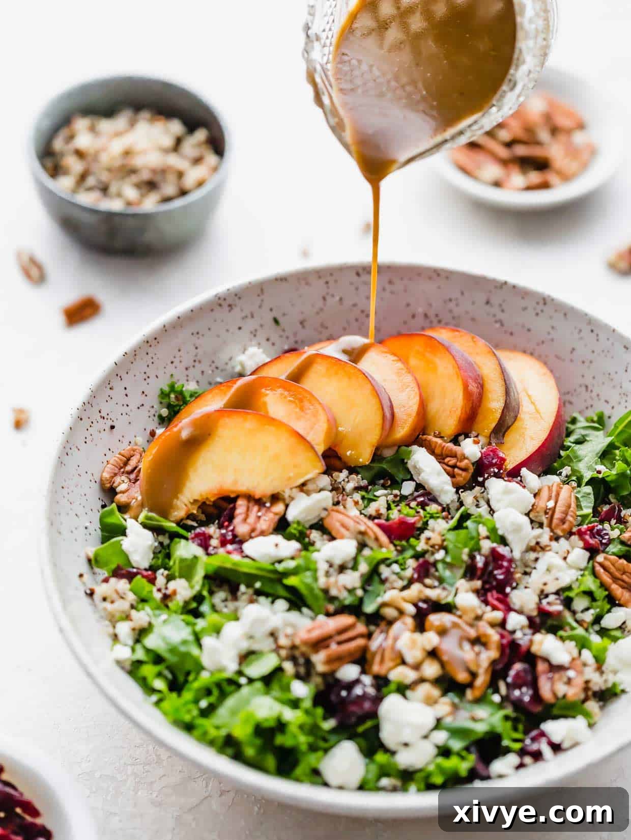 Balsamic dressing being poured overtop a kale and quinoa salad with nuts, raisins, feta cheese and sliced peaches.